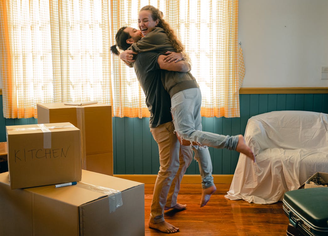 Free Side view full body barefoot young bearded male in casual clothes standing against window and lifting laughing girlfriend up during relocation and unpacking things from cardboard boxes Stock Photo